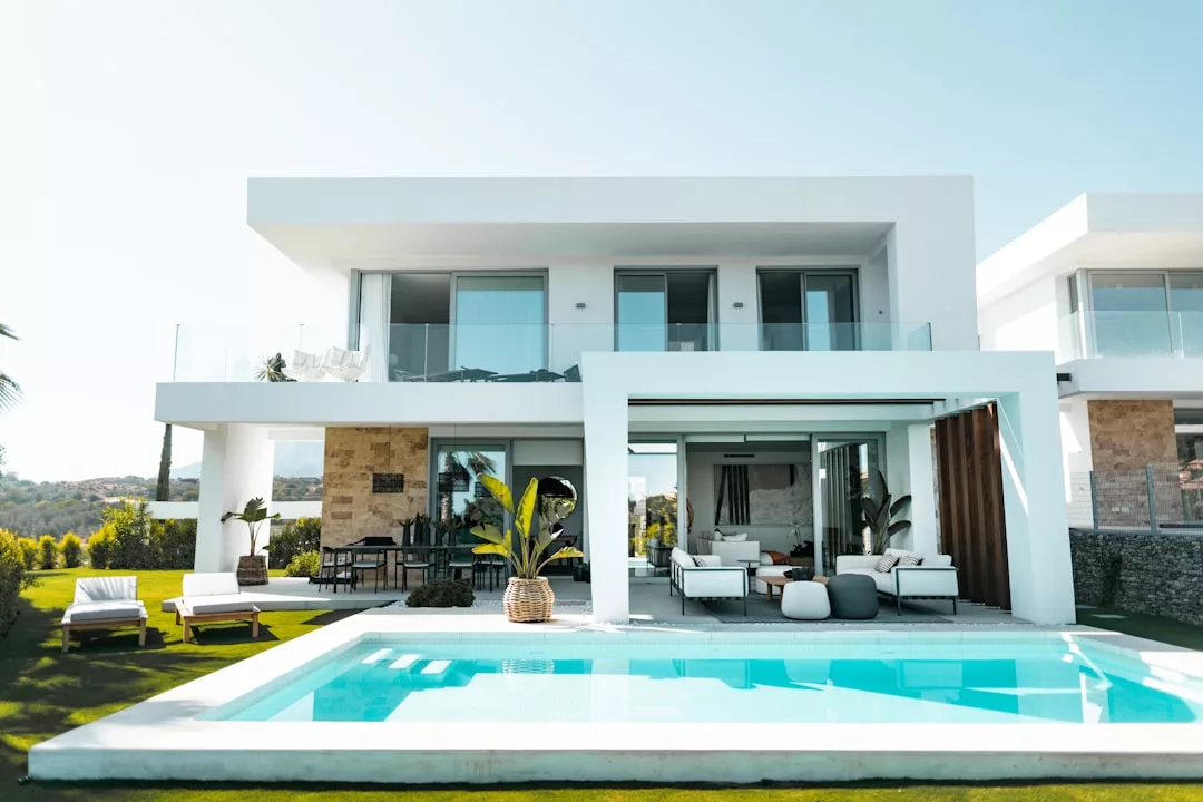 Modern two-story real estate property with large glass windows, outdoor lounge area, patio furniture, and a rectangular swimming pool in the foreground.