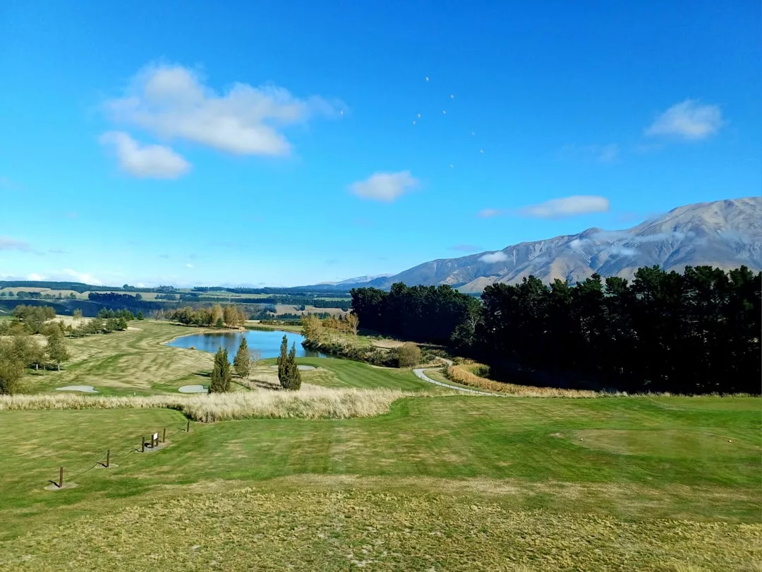 A wide view of a golf course with a pond, trees, and mountains in the background under a blue sky with scattered clouds—perfect for those seeking serene real estate surrounded by nature.
