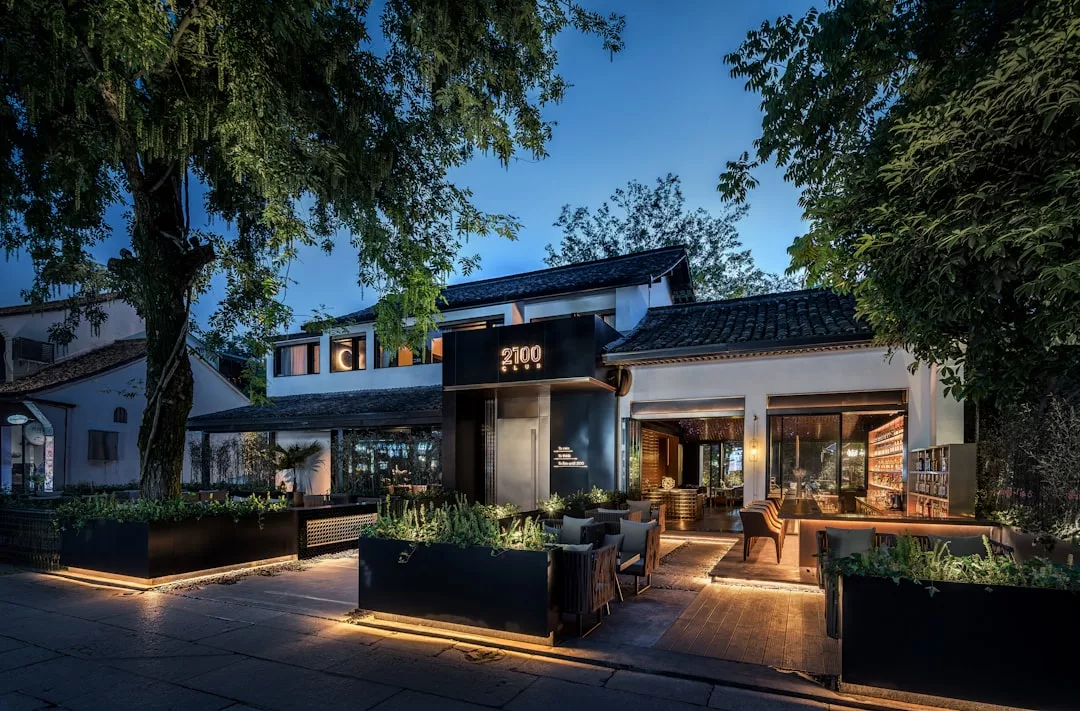 Modern restaurant exterior at dusk, featuring large windows, outdoor seating, and illuminated pathways—an inviting real estate gem nestled among trees and lush greenery.