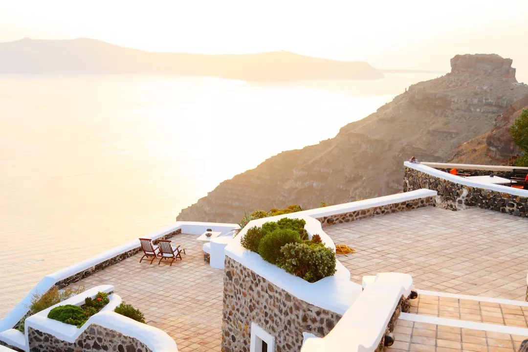 Stone terrace with white walls overlooks a calm sea and distant hills at sunset, featuring a pair of wooden lounge chairs, potted plants, and exquisite real estate perfect for tranquil living.