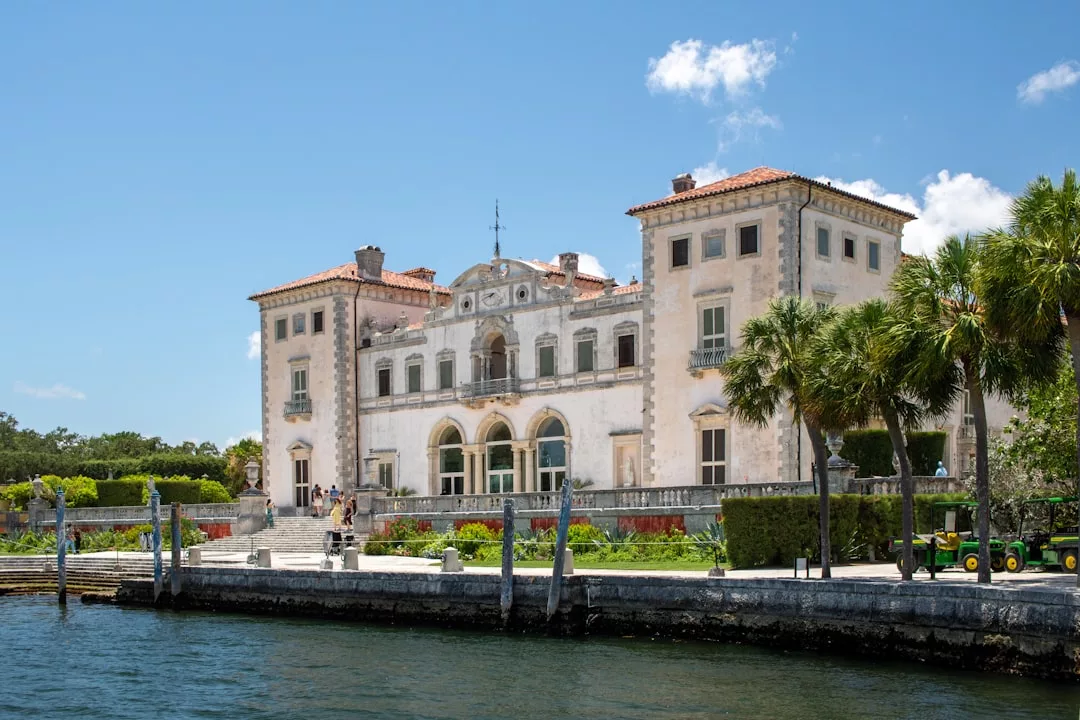 A large, historic mansion with arched windows and a red-tiled roof sits by the water, surrounded by palm trees and greenery under a clear blue sky—a true Real Estate gem.