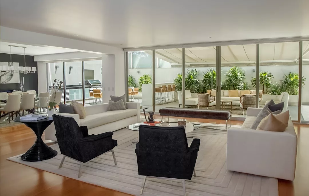 Modern living room with white sofas, black armchairs, a glass coffee table, and large sliding glass doors opening to a patio with greenery—perfect for stylish real estate showings and comfortable outdoor living.