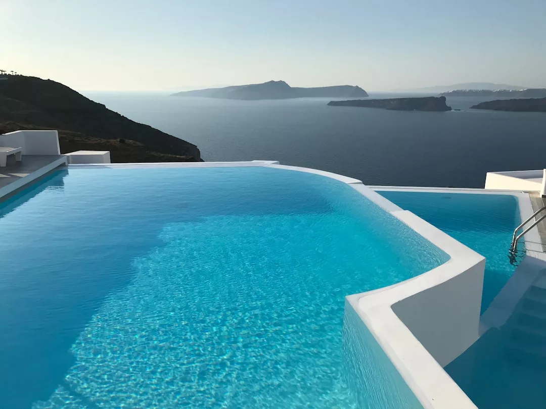 Infinity pool with clear blue water overlooks the ocean and distant islands under a clear sky, with elegant white Real Estate structures surrounding the pool.