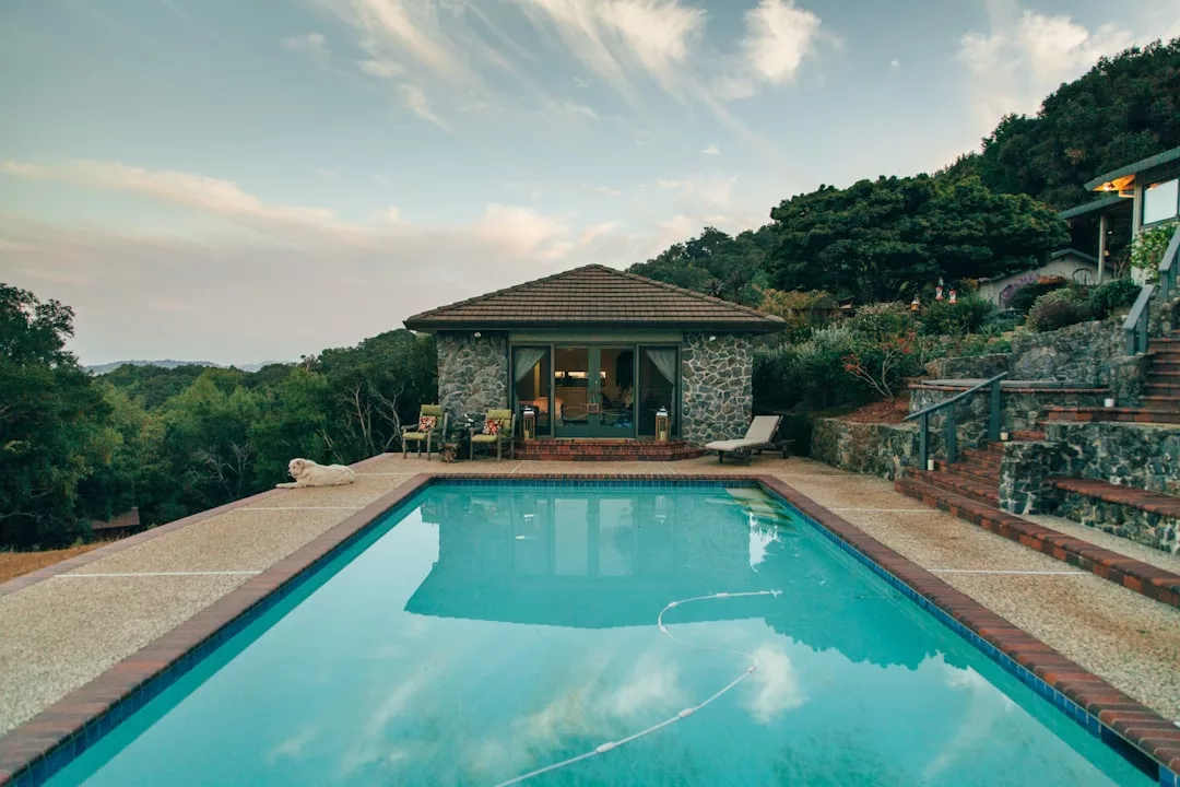 A rectangular outdoor swimming pool, ideal for real estate showcase, is bordered by stone pavement and faces a small stone building with glass doors, surrounded by trees and hills under a partly cloudy sky.