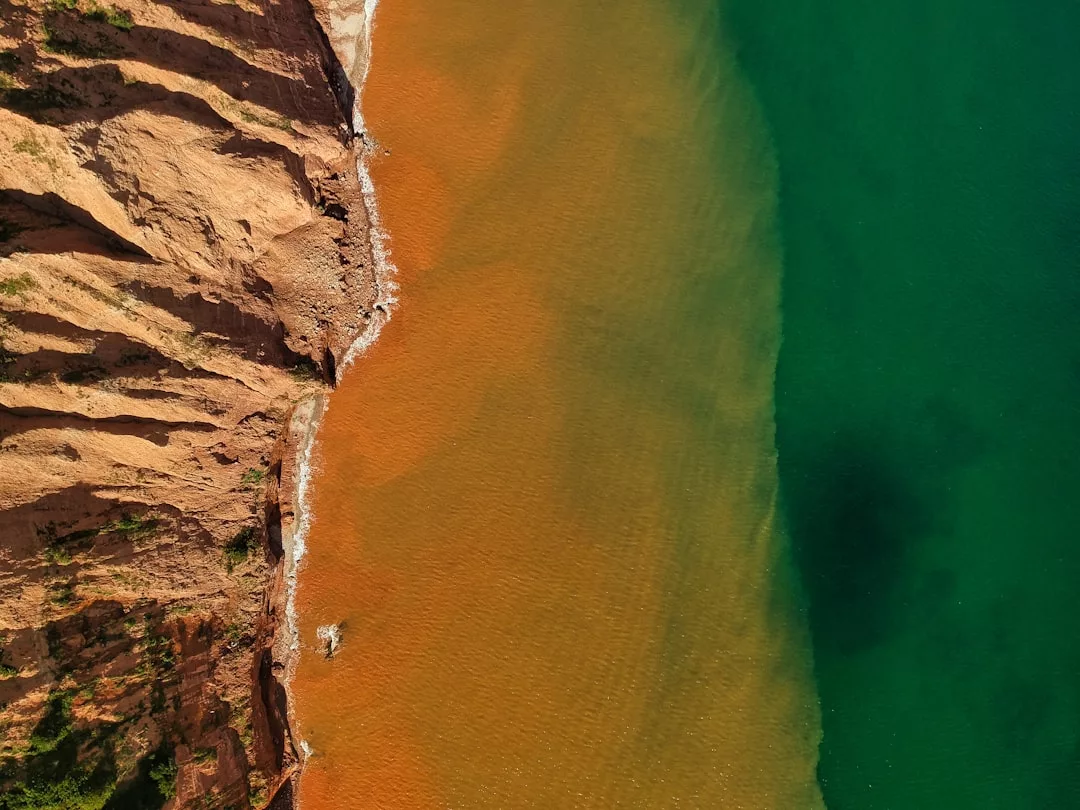 Aerial view of a reddish-brown cliff meeting a shoreline, with orange and green water blending at the edge of the land—an exceptional real estate opportunity offering stunning natural beauty.