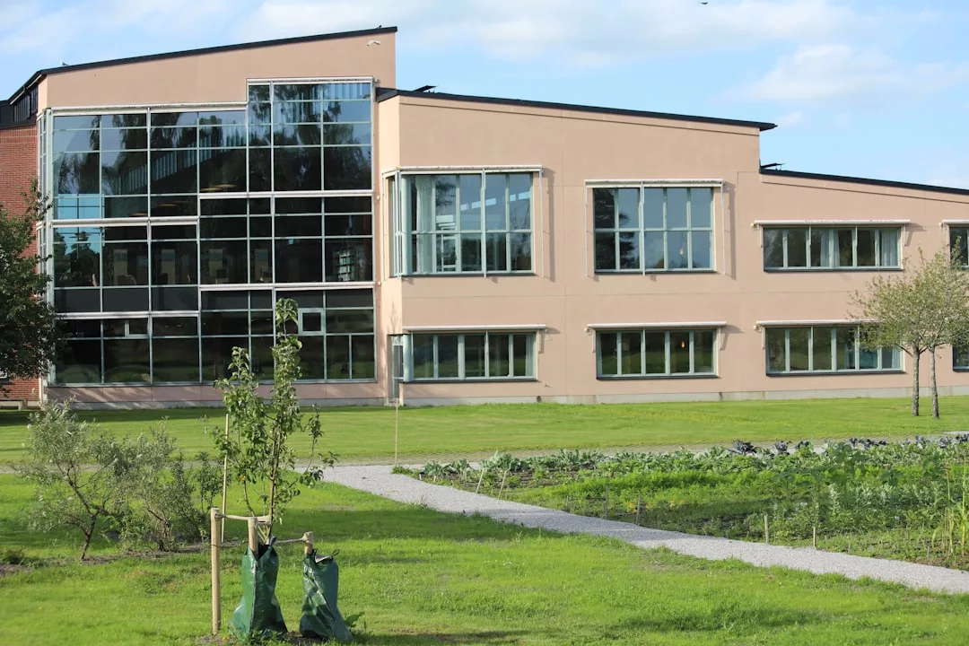 Modern two-story real estate building with large glass windows, surrounded by green lawns, young trees, and a gravel path under a partly cloudy sky.