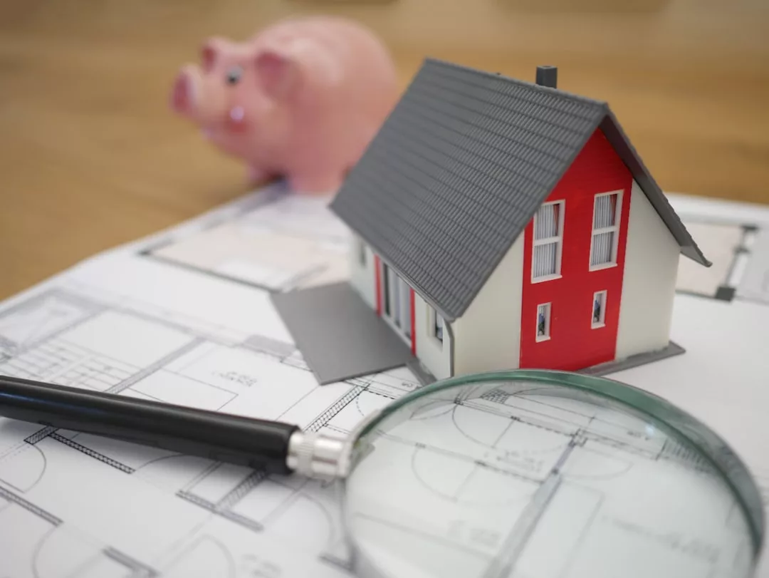A model house, magnifying glass, and pen rest on architectural blueprints, with a pink piggy bank in the background—perfectly capturing the essence of real estate planning.