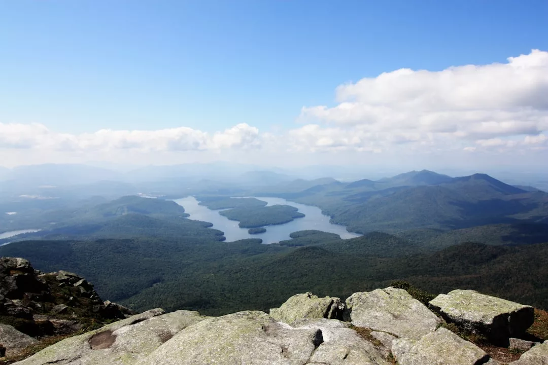 A view from a rocky mountaintop overlooks a winding lake surrounded by forested hills under a partly cloudy sky, offering real estate seekers breathtaking natural beauty and serene landscapes.