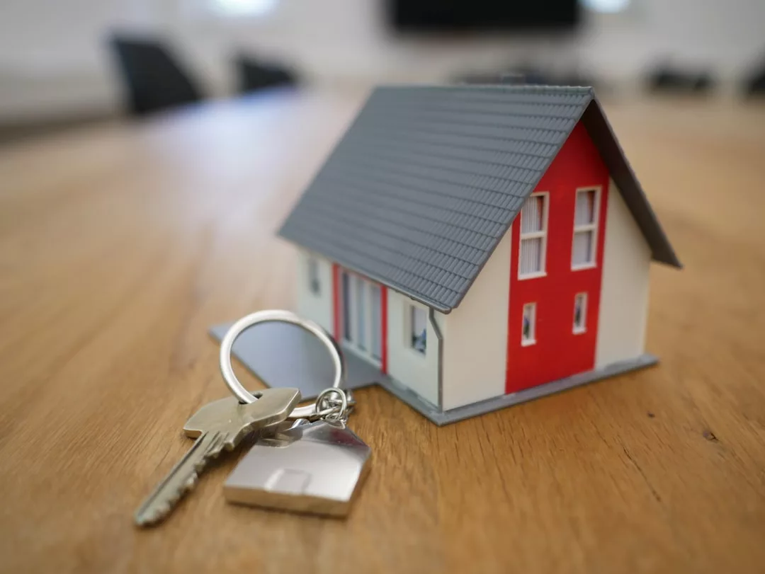 A small model house sits on a wooden table next to a set of keys with a house-shaped keychain, symbolizing new beginnings in real estate.