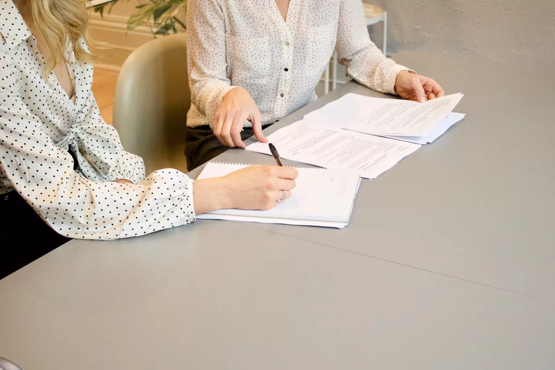 Two people in polka dot blouses sit at a table reviewing and marking real estate documents with pens.