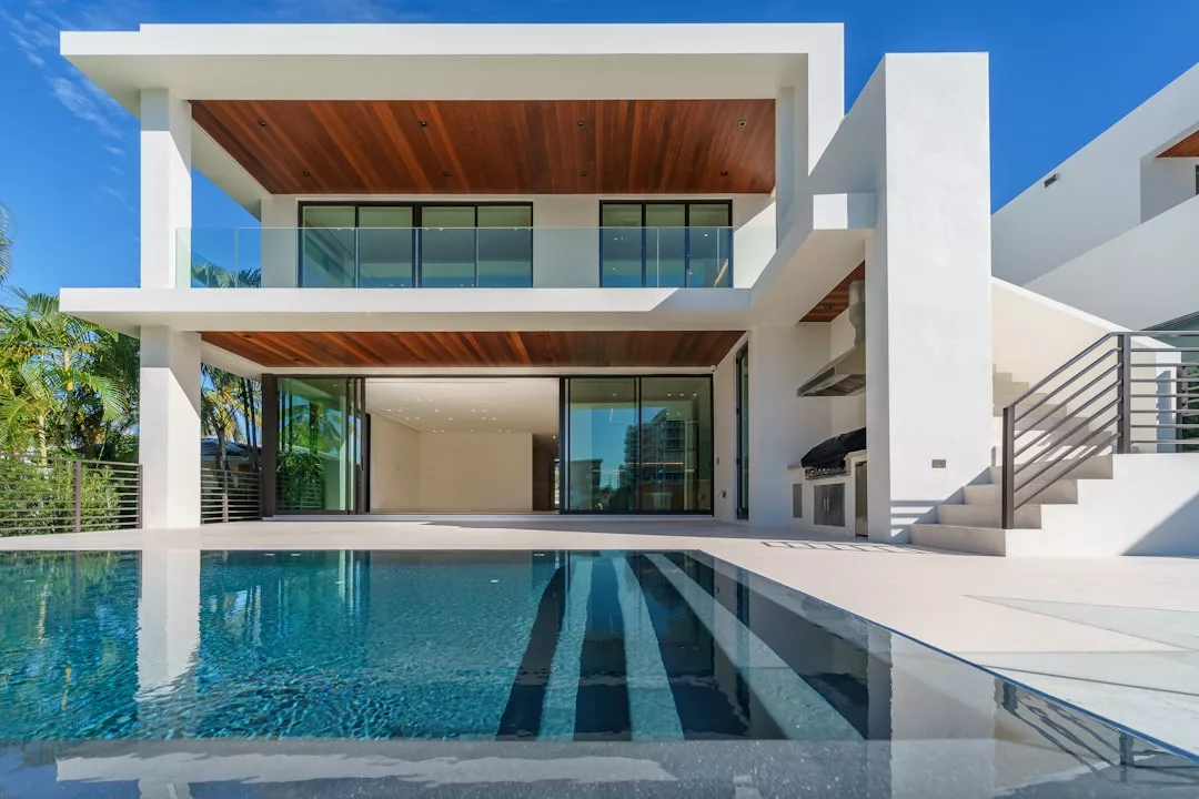 Modern two-story Real Estate property with large glass windows, wooden ceilings, a pool in the foreground, and a built-in outdoor grill under a covered patio.