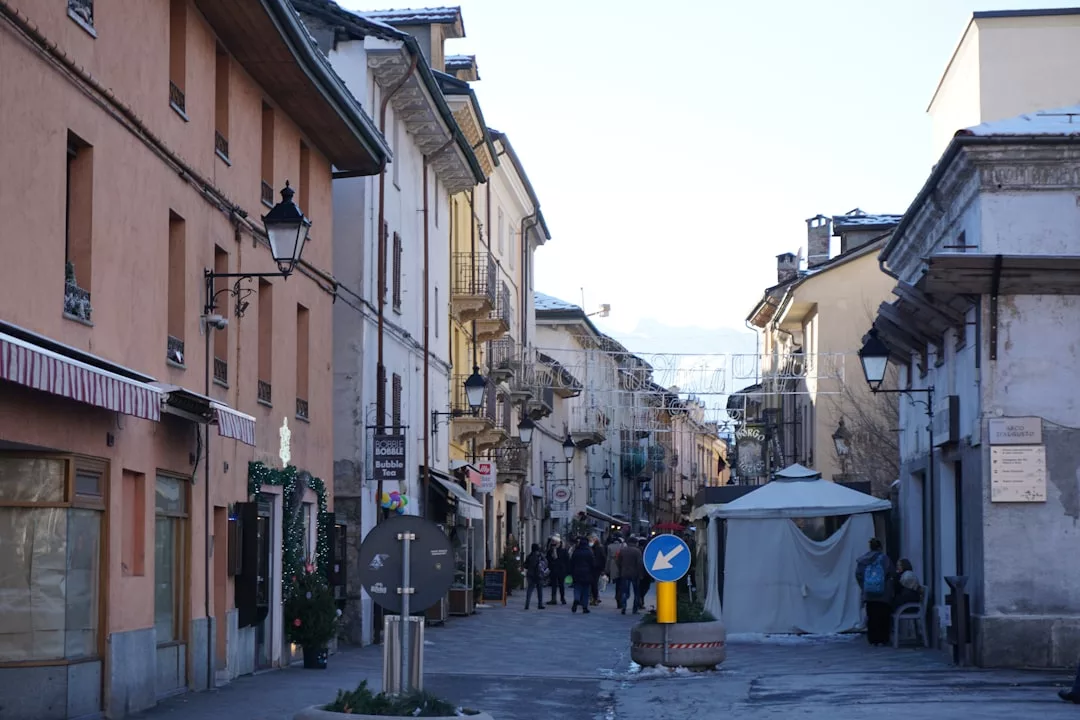 A narrow street in a European town with charming buildings on both sides, people walking, a tent structure, and a blue roundabout traffic sign—an inviting scene perfect for those seeking unique real estate opportunities.