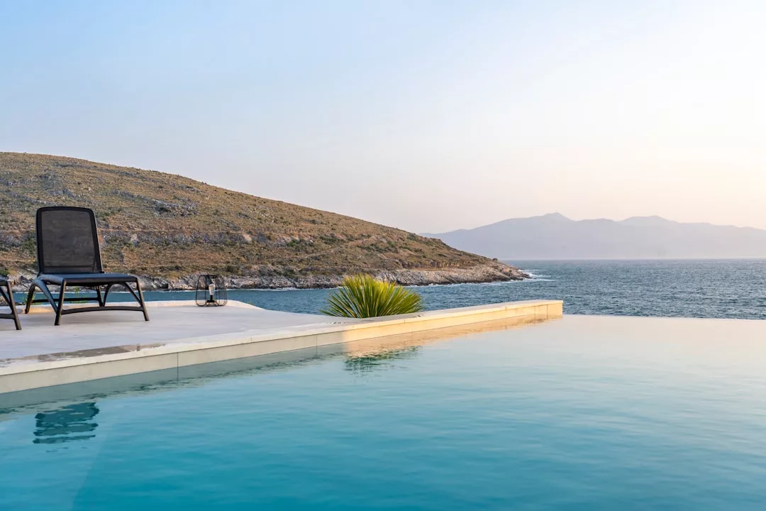 A single lounge chair sits by an infinity pool overlooking the sea—a stunning real estate setting with hills and mountains visible in the background under a clear sky.