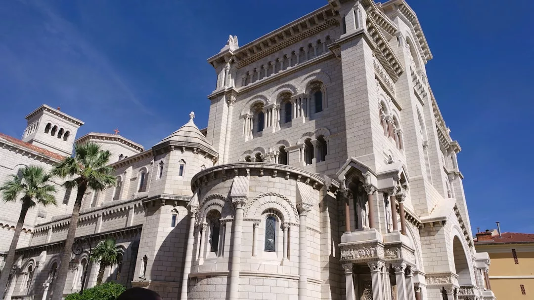 Large stone cathedral with arched windows and detailed carvings, set against a clear blue sky, surrounded by palm trees and nearby real estate buildings.