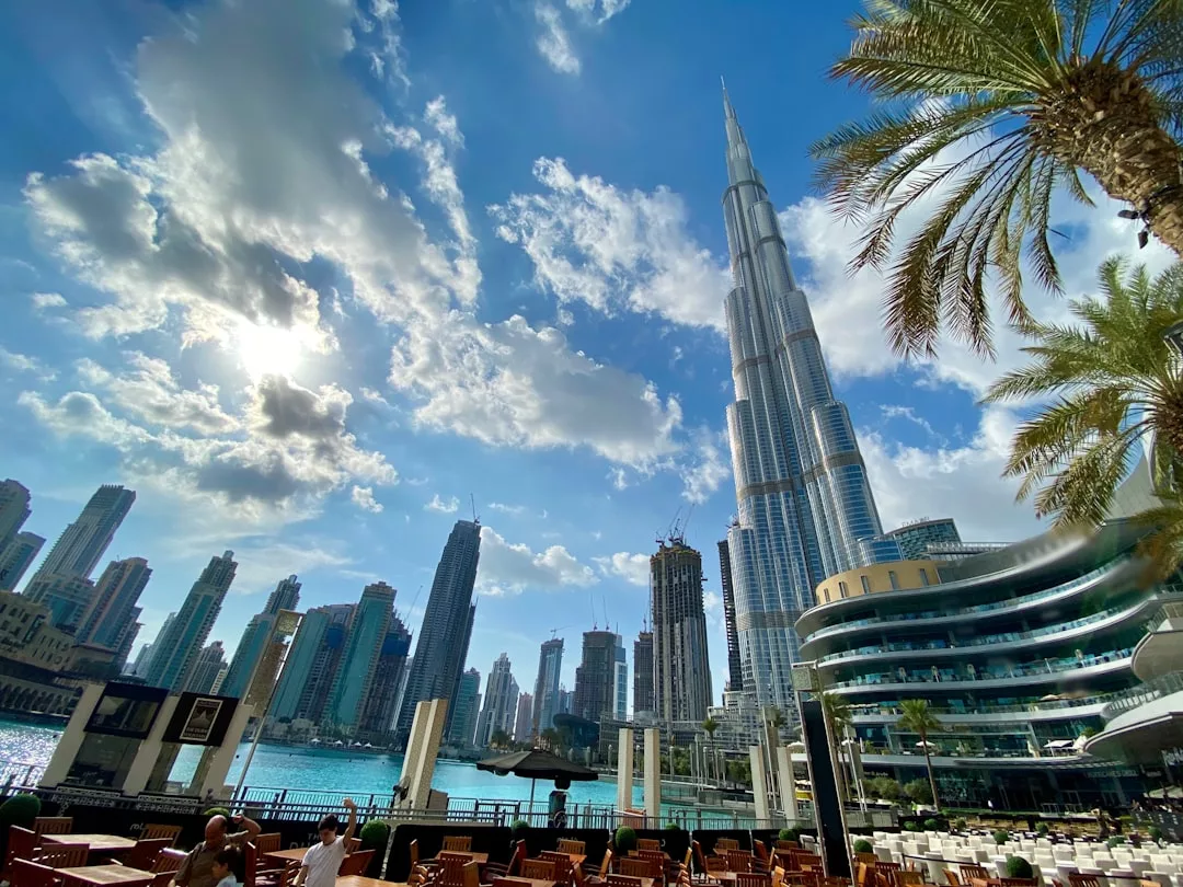Outdoor restaurant seating in Dubai with the Burj Khalifa and surrounding skyscrapers, showcasing stunning real estate under a partly cloudy sky and palm trees in the foreground.