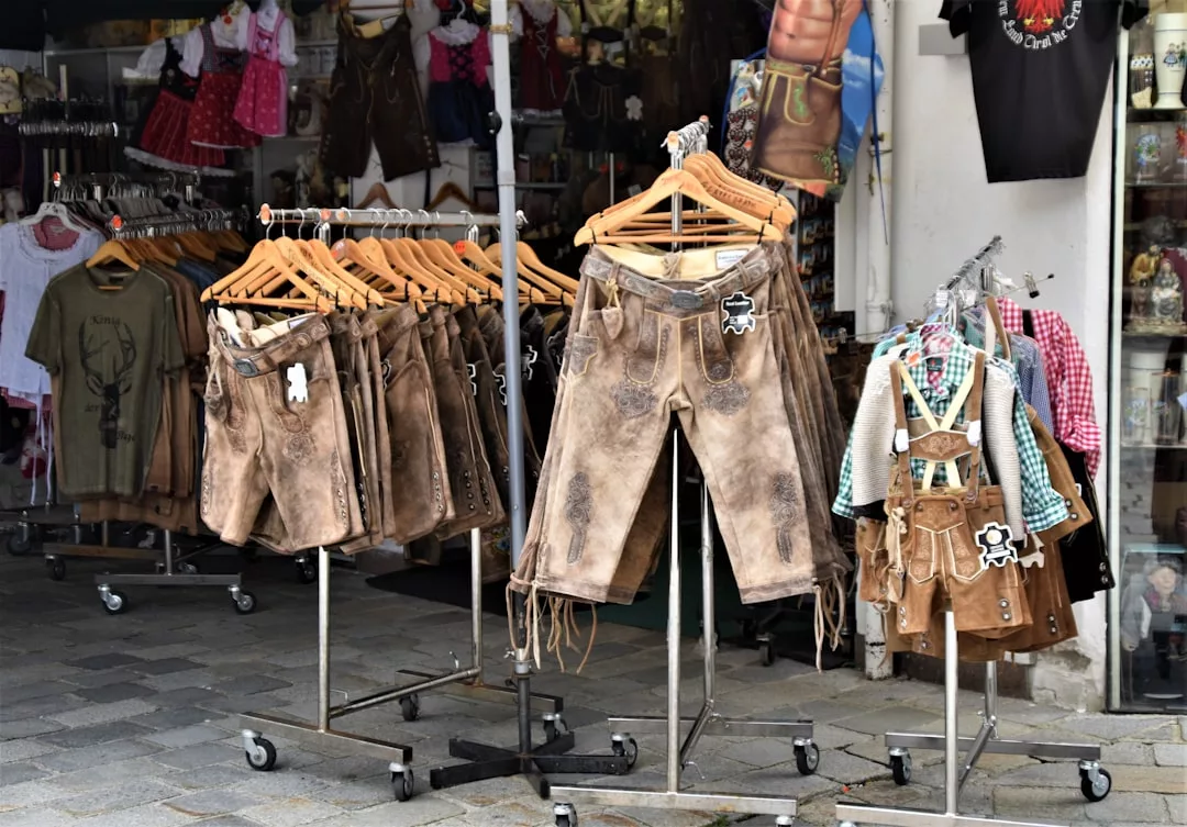 Several racks of traditional brown leather lederhosen and shirts are displayed outside a shop, with more traditional clothing visible inside—reflecting the region’s rich heritage and timeless appeal that draws both tourists and real estate investors alike.