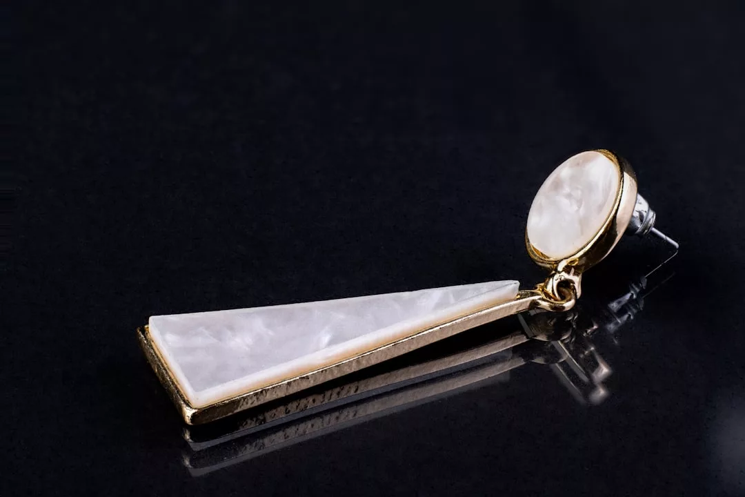 A single gold and white earring with a round stud and a long, triangular drop design is showcased on a black surface, reflecting elegance akin to exquisite real estate.