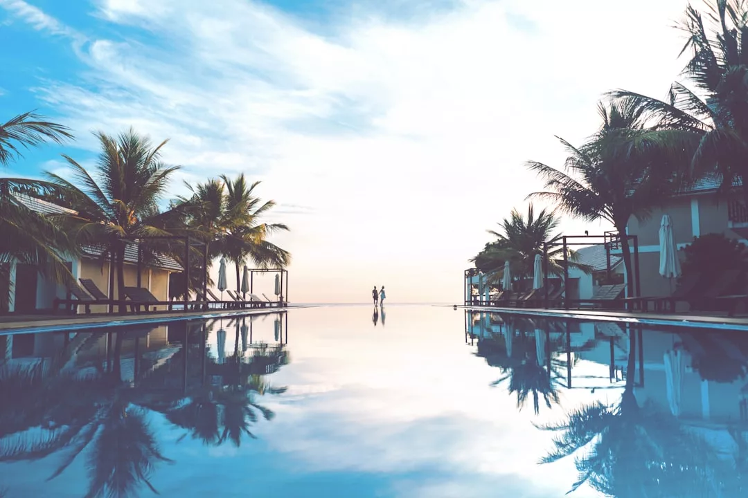 A large, calm swimming pool is flanked by palm trees and lounge chairs, offering a serene real estate retreat, with two people standing in the distance at the water’s edge under a partly cloudy sky.
