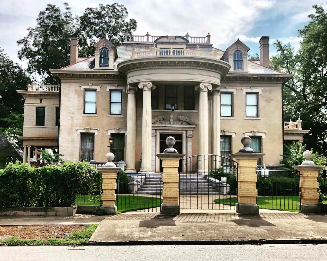 A large, two-story historic mansion with tall columns at the entrance, ornate architectural details, and a gated front yard surrounded by greenery—an exceptional real estate opportunity.