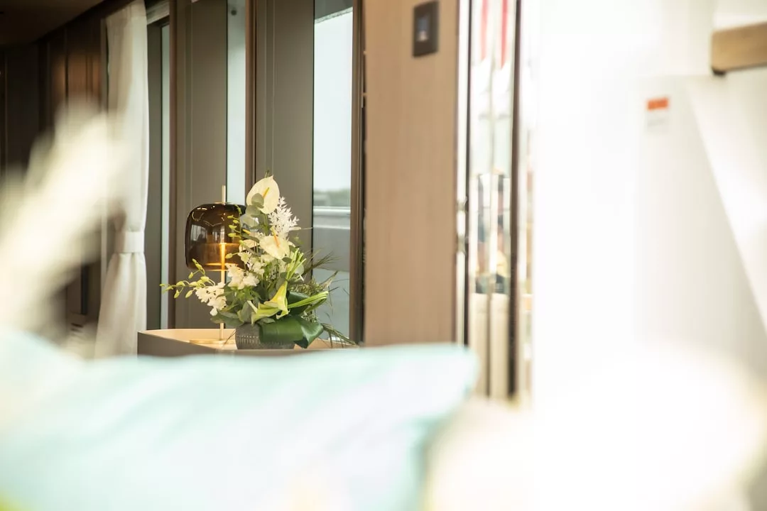 A modern interior in a real estate setting features a glass lamp and floral arrangement on a table near large windows, filling the space with natural light.