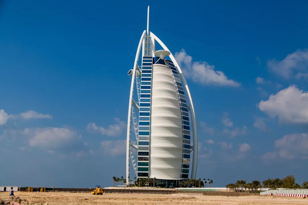The Burj Al Arab hotel in Dubai, an icon of modern real estate, rises like a sail-shaped skyscraper against a blue sky with scattered clouds and a sandy foreground.