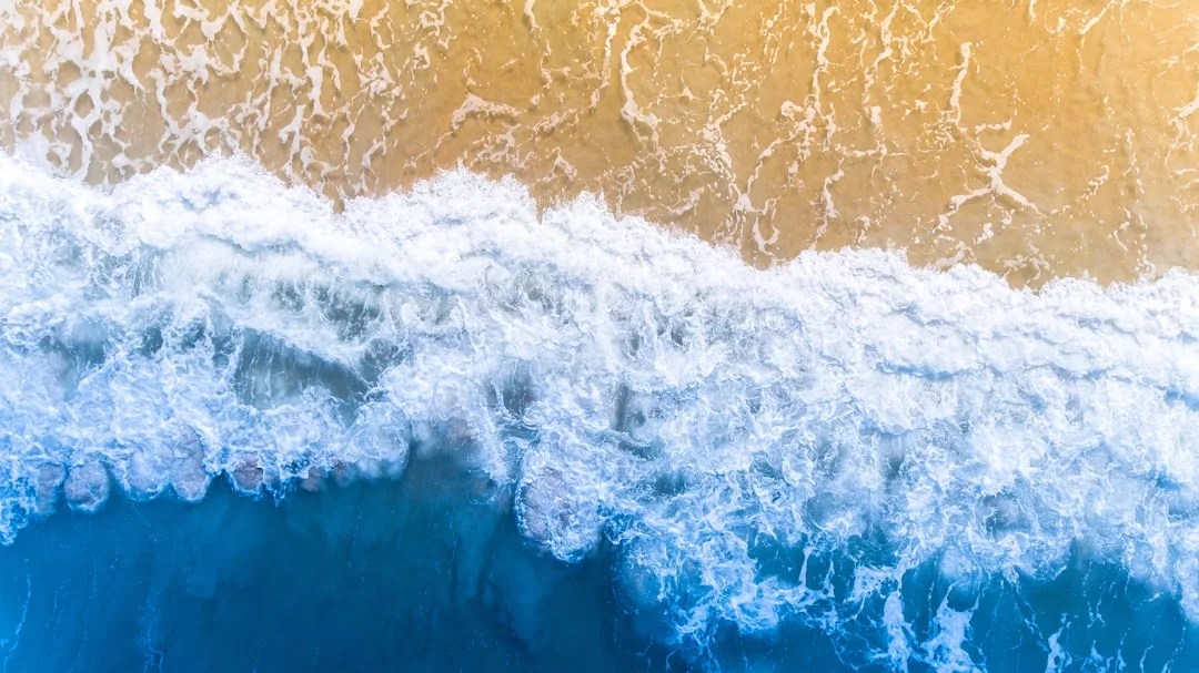 Aerial view of ocean waves crashing onto a sandy beach, with white foam separating blue water from tan sand—a stunning real estate perspective of coastal beauty.