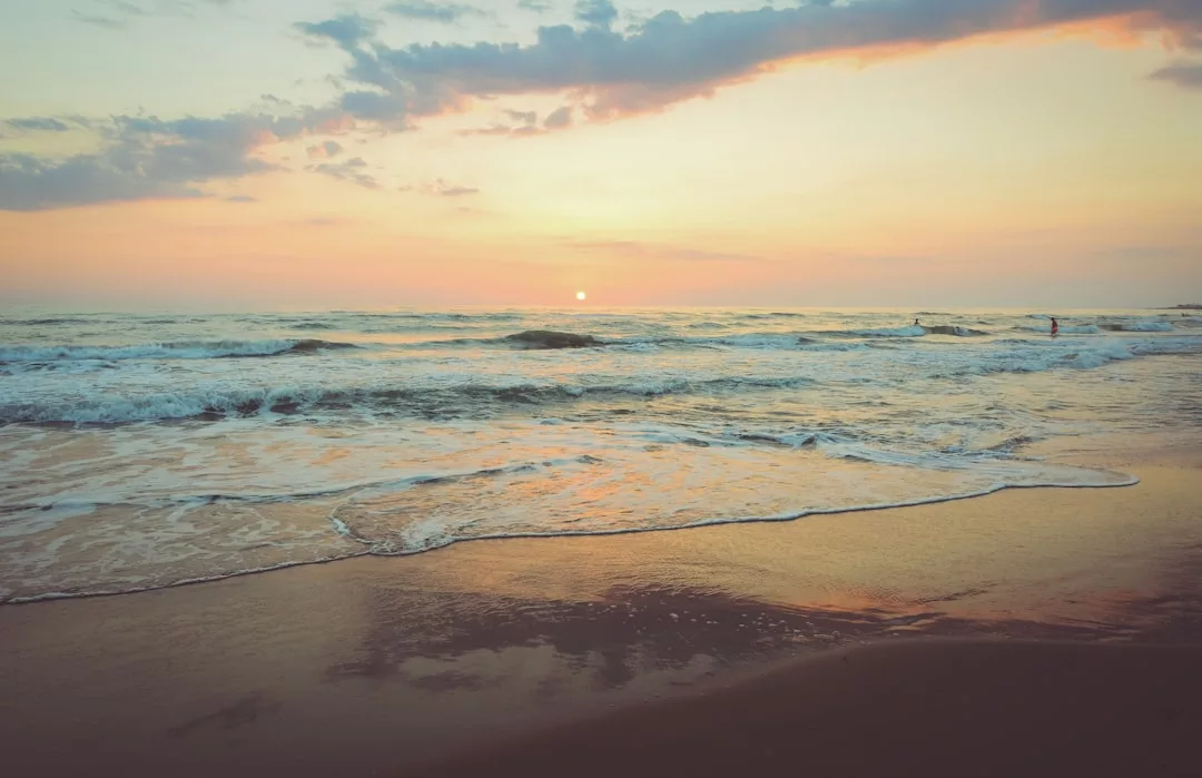A calm beach with gentle waves and a pastel-colored sky at sunrise or sunset creates an inviting backdrop, perfect for real estate that offers serene ocean views; the sun is low on the horizon with clouds scattered above the water.