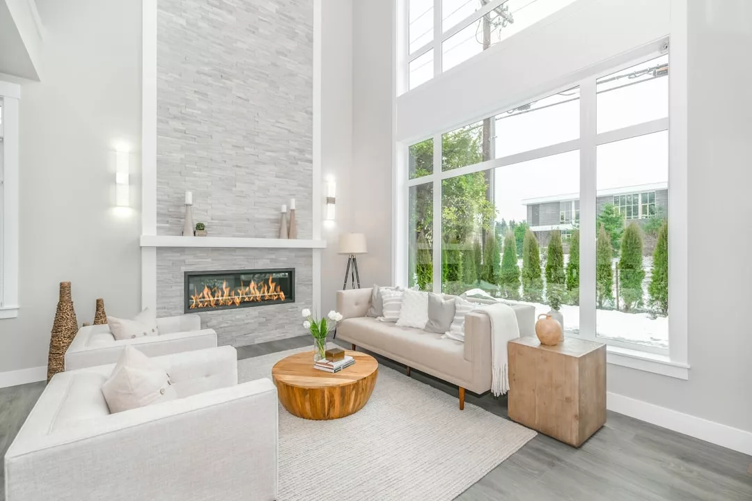 Bright modern living room with tall windows, light-colored furniture, a stone fireplace, wooden coffee table, and decorative elements on a neutral rug—perfect for showcasing the best in real estate.
