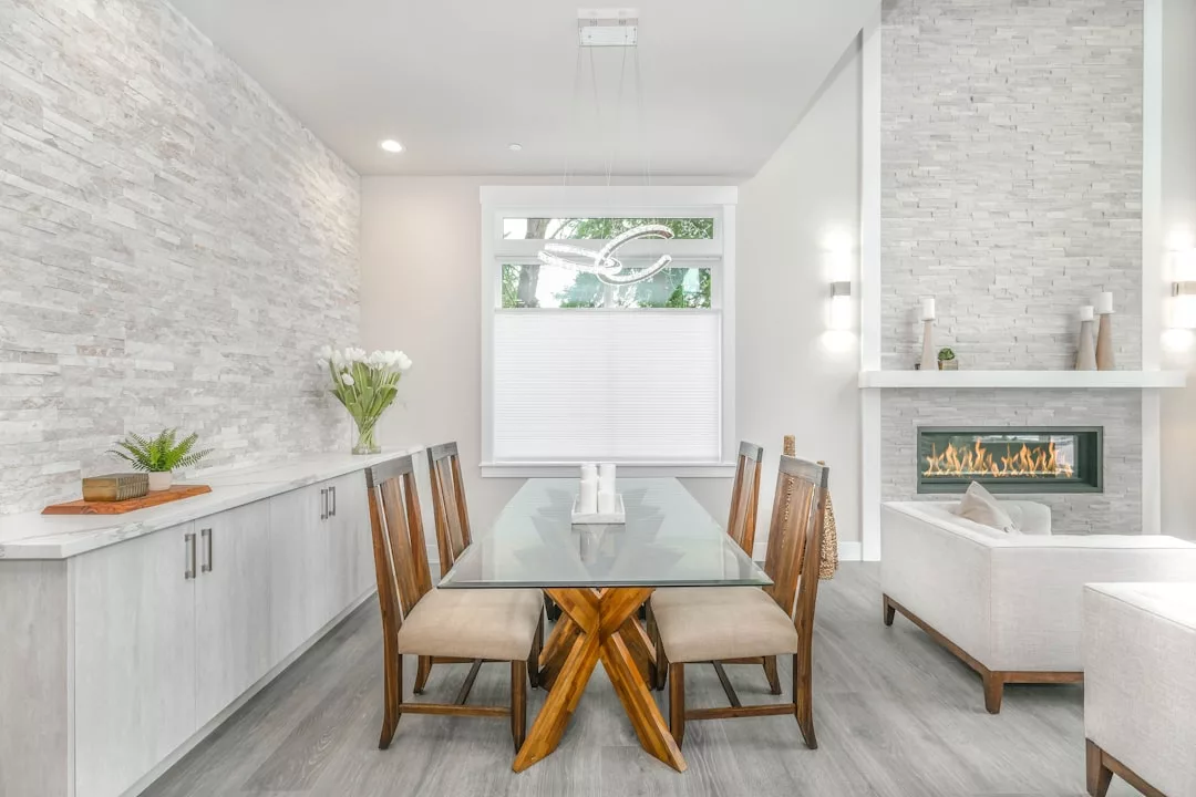 Modern dining room in this stunning real estate features a glass table, wooden chairs, light stone accent walls, built-in cabinets, a large window, and a contemporary fireplace in the adjacent living area.