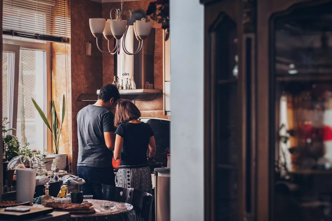 Two people stand side by side in a kitchen, preparing food together at the counter, surrounded by various items and plants—a warm scene that highlights the inviting atmosphere of this real estate gem.