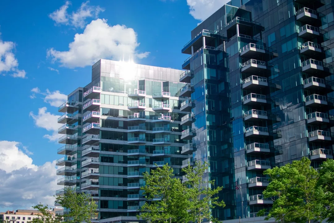 Modern glass apartment buildings with balconies reflect sunlight against a blue sky with clouds, with green trees in the foreground—an inviting scene for discerning real estate seekers.