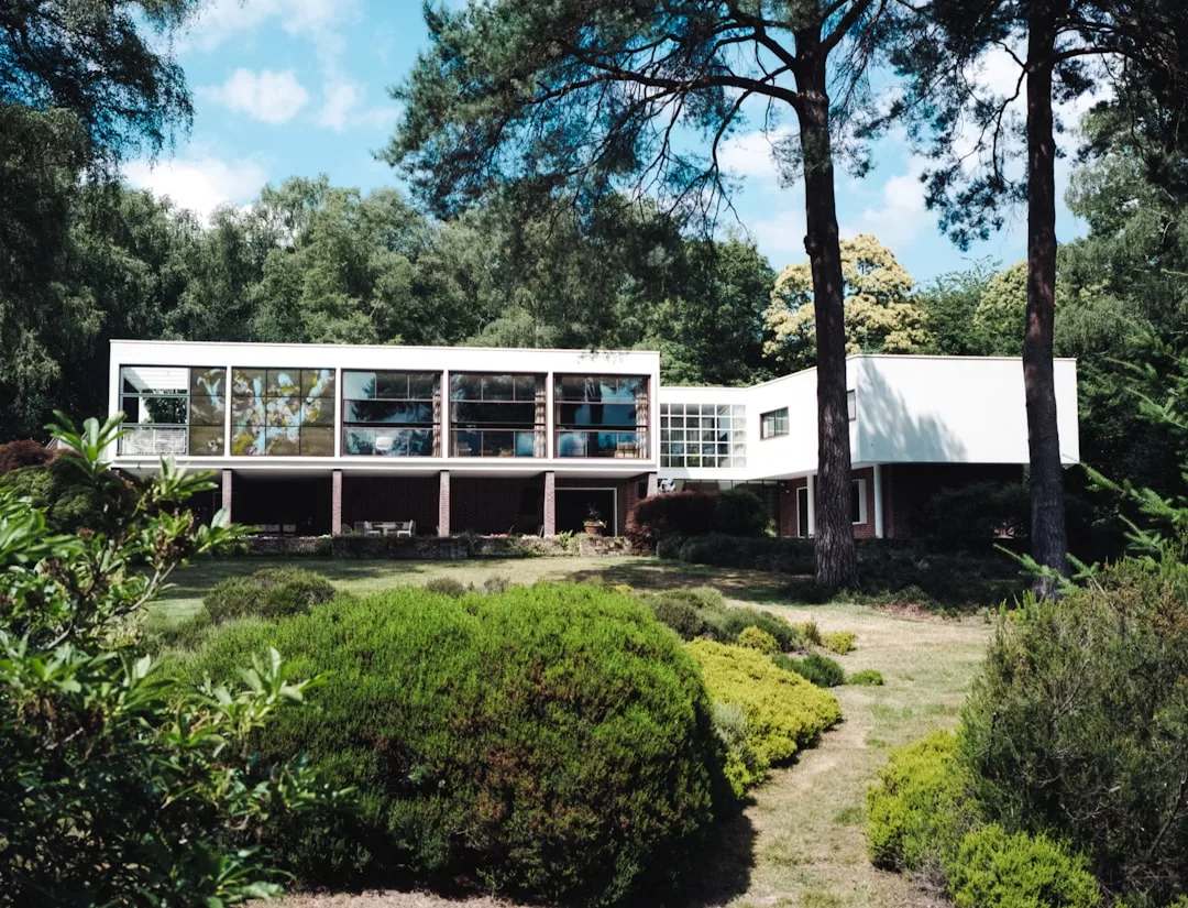 A modern two-story Real Estate property with large windows and flat roofs, surrounded by trees and landscaped greenery.
