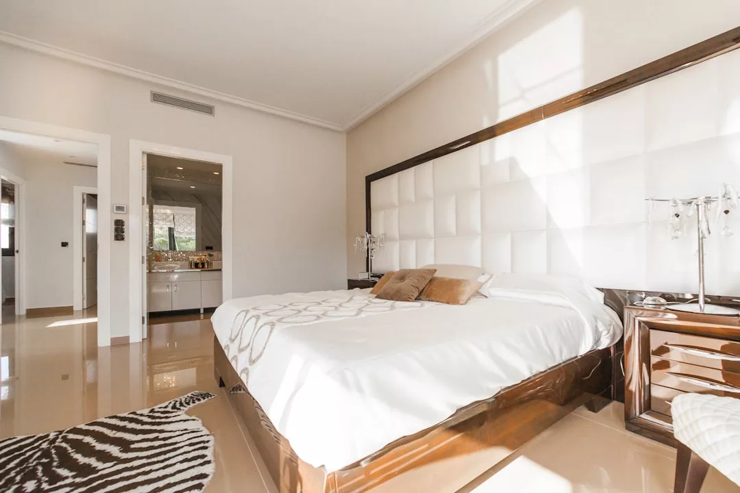 Modern bedroom designed for real estate appeal, featuring a large bed with padded headboard, bedside tables, zebra-patterned rug, and an open view into a bright bathroom area.