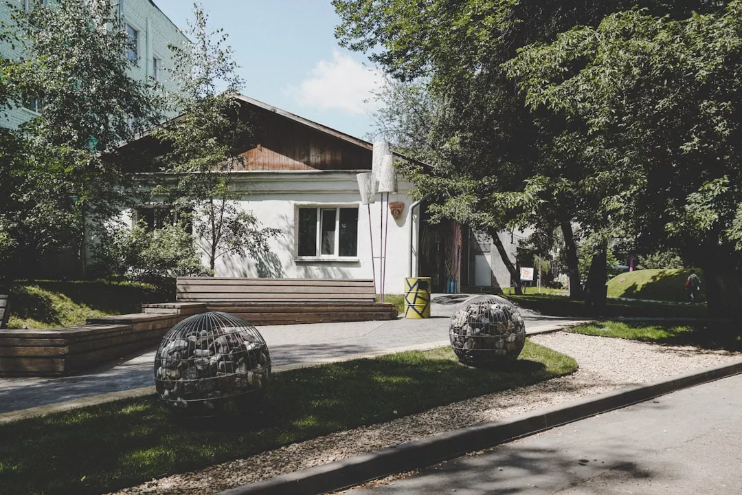 A charming small white building with a wooden roof stands behind benches, trees, and spherical outdoor sculptures—perfect for unique real estate opportunities on a sunny day.