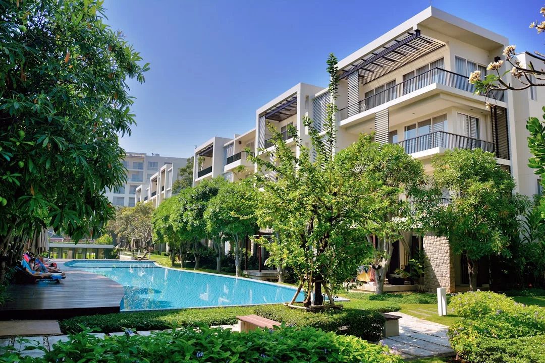 Modern real estate apartment buildings surround a rectangular outdoor swimming pool, with trees and greenery around the area under a clear blue sky.