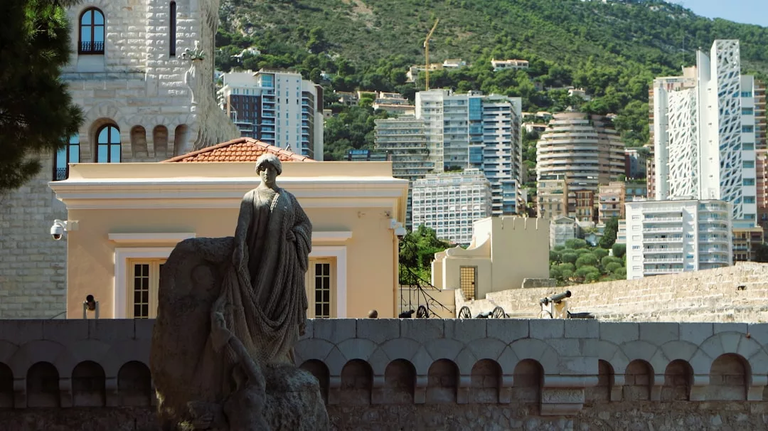 A stone statue stands in the foreground near a low stone wall, with modern real estate high-rise buildings and green hills in the background.