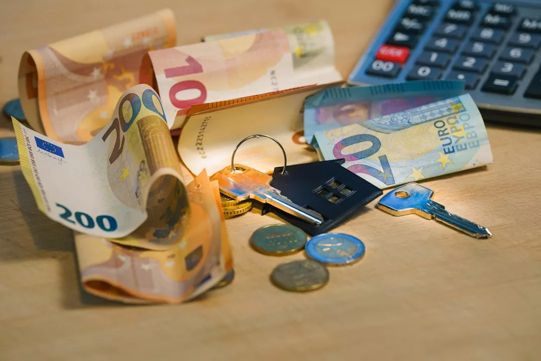 Euro banknotes, coins, two keys with a house-shaped keychain, and a calculator are placed on a wooden surface—elements often associated with Real Estate transactions.