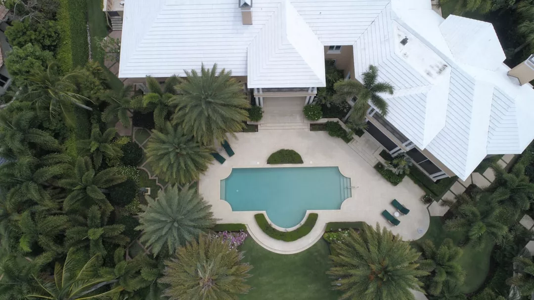 Aerial view of a Real Estate property with a white-roofed house, surrounded by palm trees, featuring a rectangular swimming pool and lounge chairs in the backyard.
