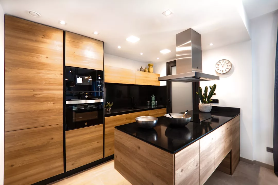 Modern kitchen with wood cabinetry, black countertops, built-in ovens, a central island cooktop, metal vent hood, wall clock—perfect for real estate buyers seeking style and functionality.