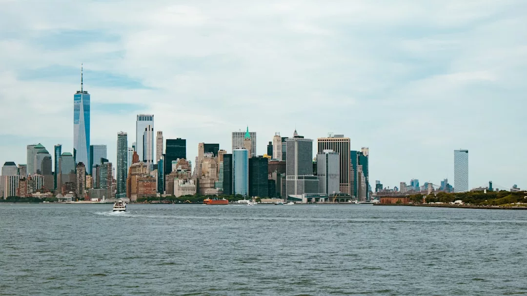 A wide view of the Manhattan skyline in New York City, showcasing tall buildings, the One World Trade Center, and a boat on the Hudson River under a cloudy sky—an iconic scene for Real Estate enthusiasts.