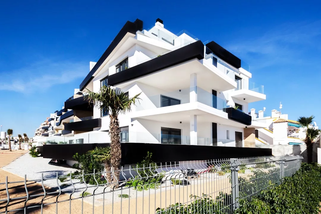Modern, multi-story white real estate building with black trim, large balconies, and glass railings, surrounded by a metal fence and palm trees under a clear blue sky.