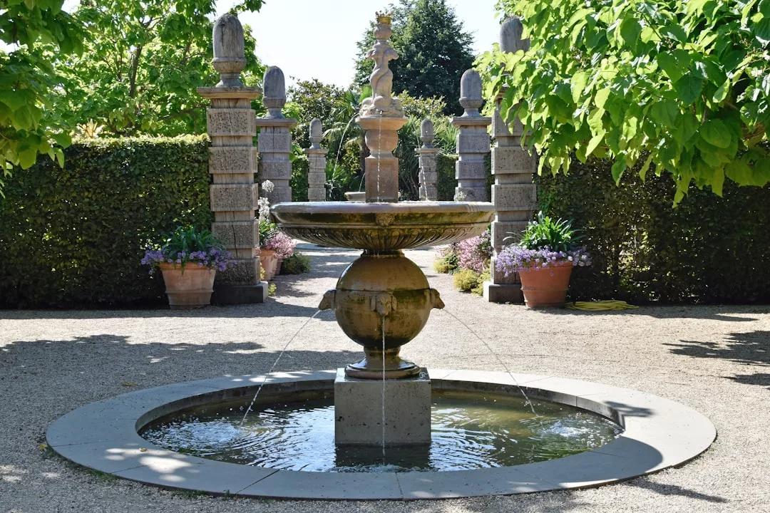 A round stone fountain with water streams stands in the center of a garden, surrounded by trees, potted plants, and stone pillars—an inviting outdoor feature perfect for enhancing your real estate’s appeal.