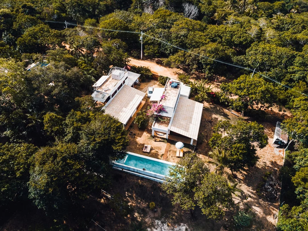 Aerial view of a modern real estate property with a rectangular pool, surrounded by dense trees and vegetation, with dirt paths and utility lines visible.