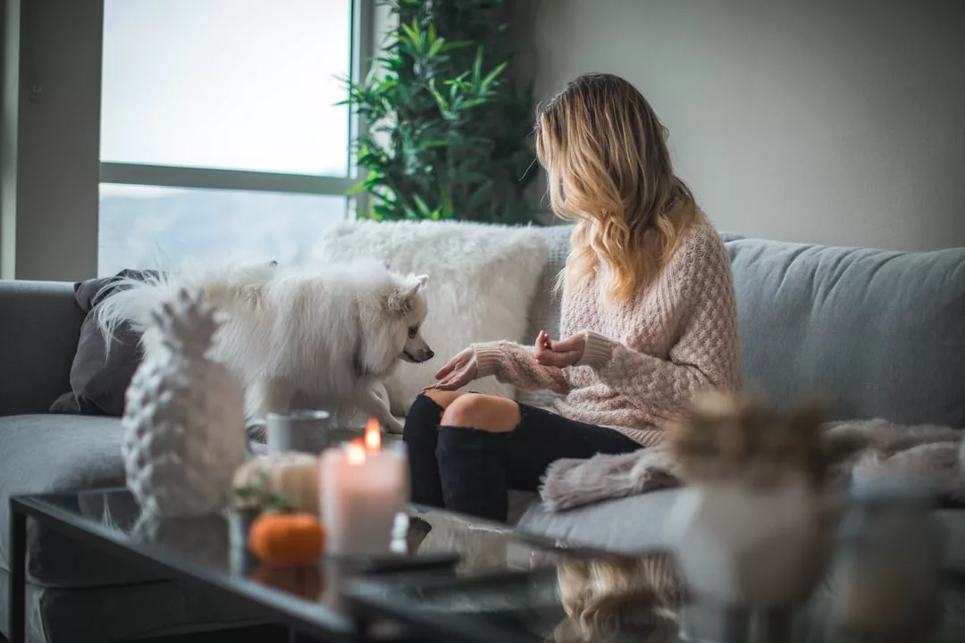 A woman sits on a gray sofa in a stylish real estate setting, holding a treat as her fluffy white dog stands beside her, with candles and decor arranged on a glass coffee table in front.