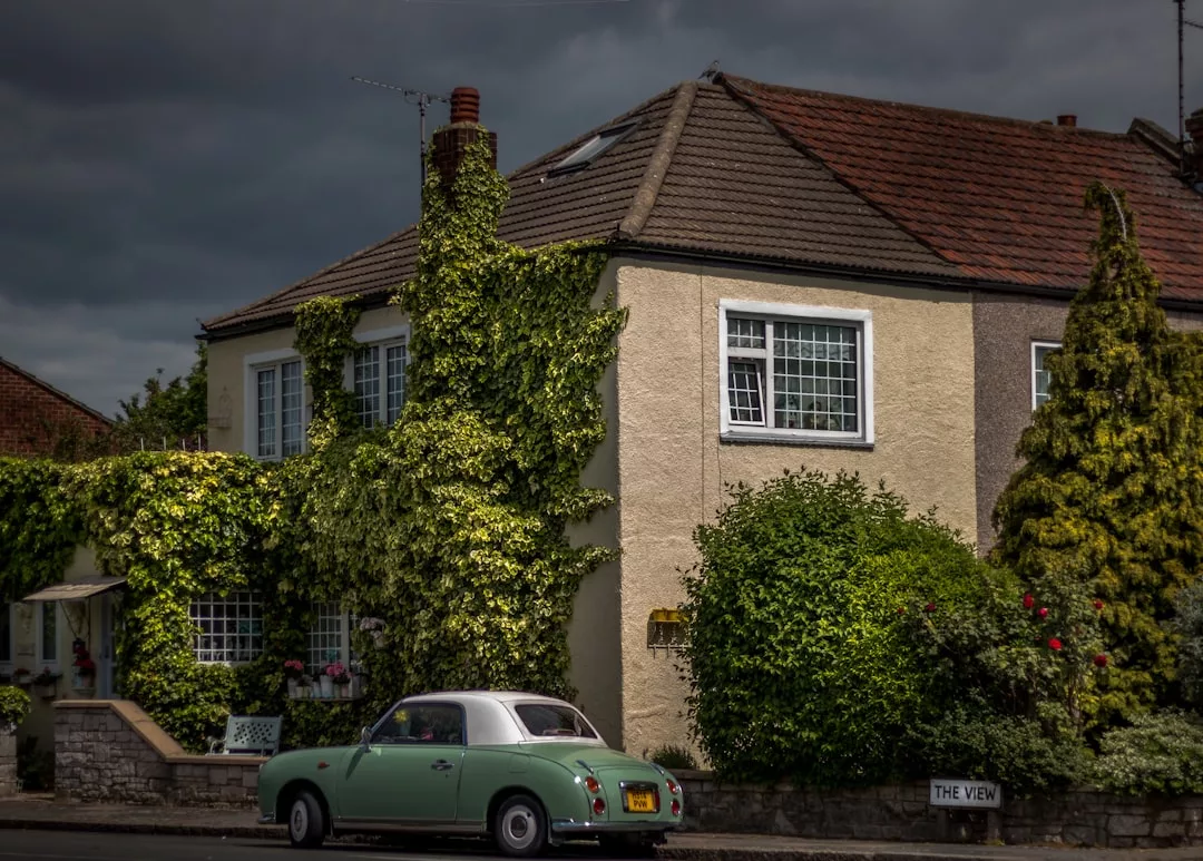 A vintage green car is parked on the street in front of a charming two-story house covered in ivy, a picturesque spot that highlights classic real estate appeal, surrounded by lush greenery under a cloudy sky.