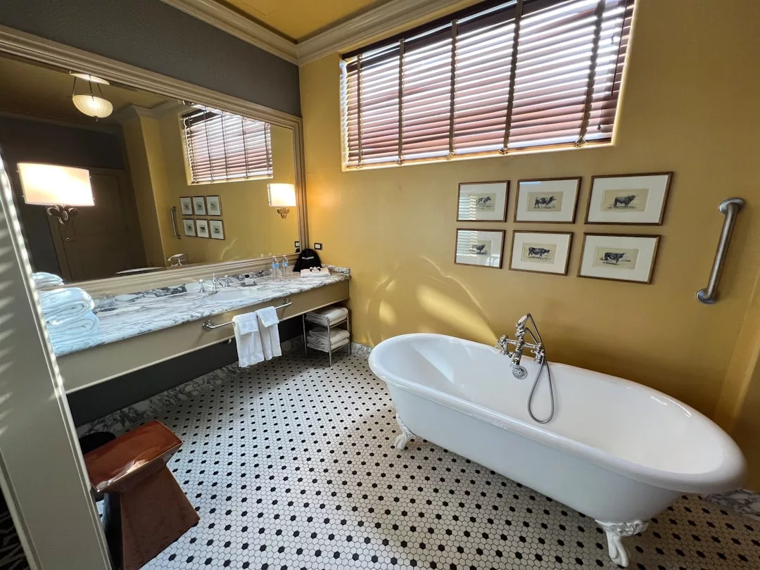 A stunning bathroom perfect for real estate listings, featuring a freestanding clawfoot bathtub, marble countertop, wall mirror, folded towels, framed pictures, and a window with blinds.
