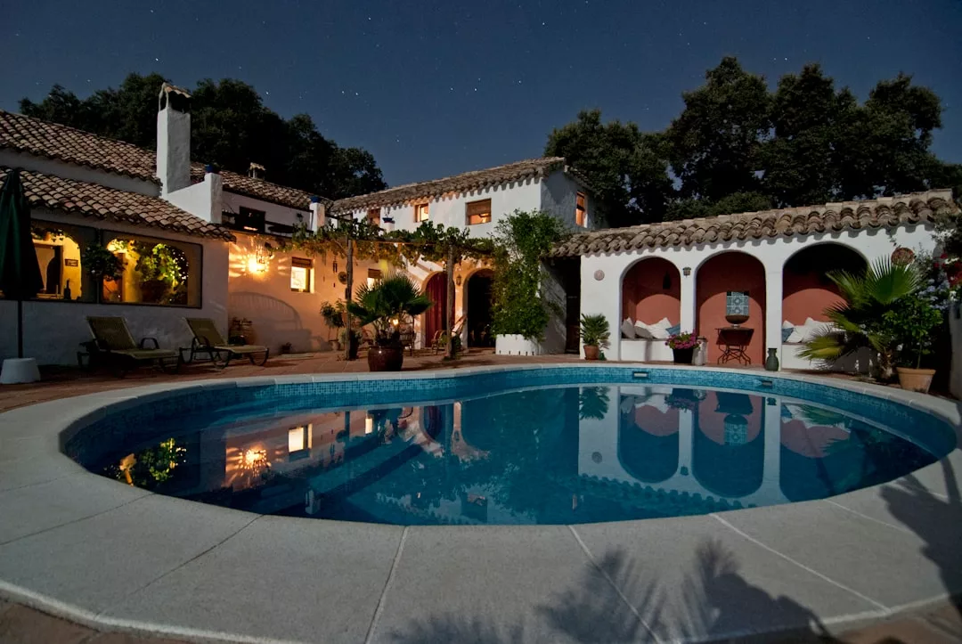 A courtyard with a circular swimming pool is surrounded by a Spanish-style real estate villa, lit by outdoor lamps at night under a clear sky.