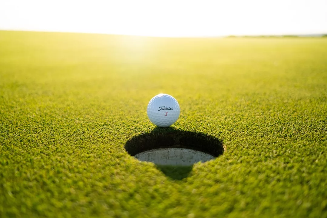 A golf ball rests on the edge of a hole on a pristine golf course green, highlighting the allure of real estate in premium golfing communities under bright sunlight.