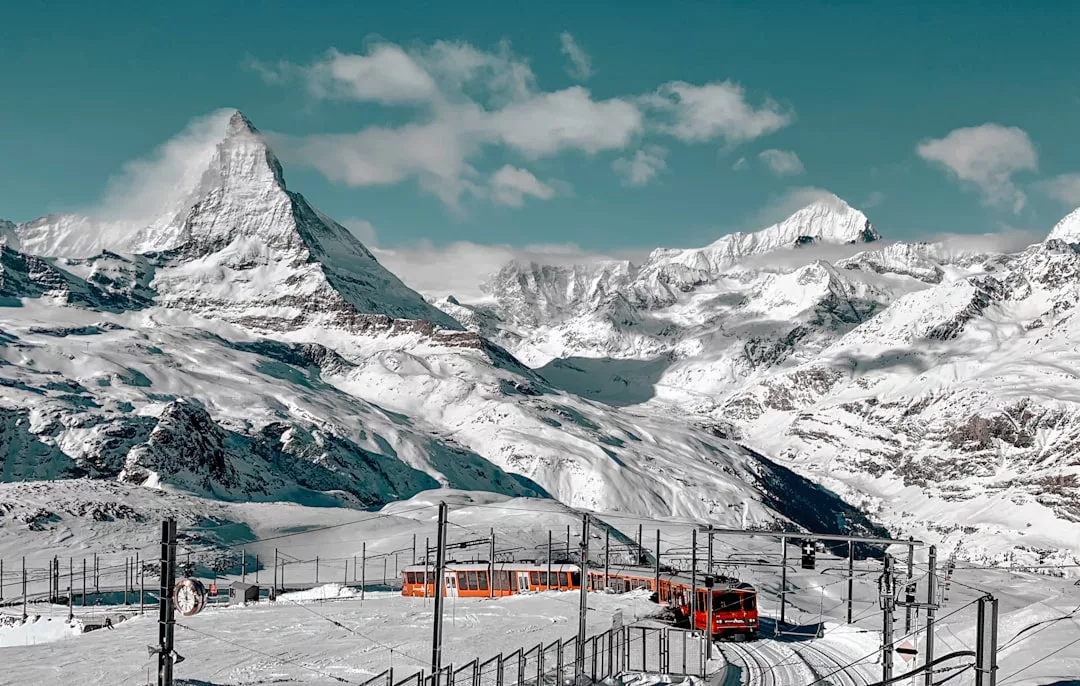 A red train travels on snowy tracks with the Matterhorn and surrounding snow-covered Alps in the background under a clear sky, showcasing the breathtaking landscape that makes Swiss real estate highly coveted.