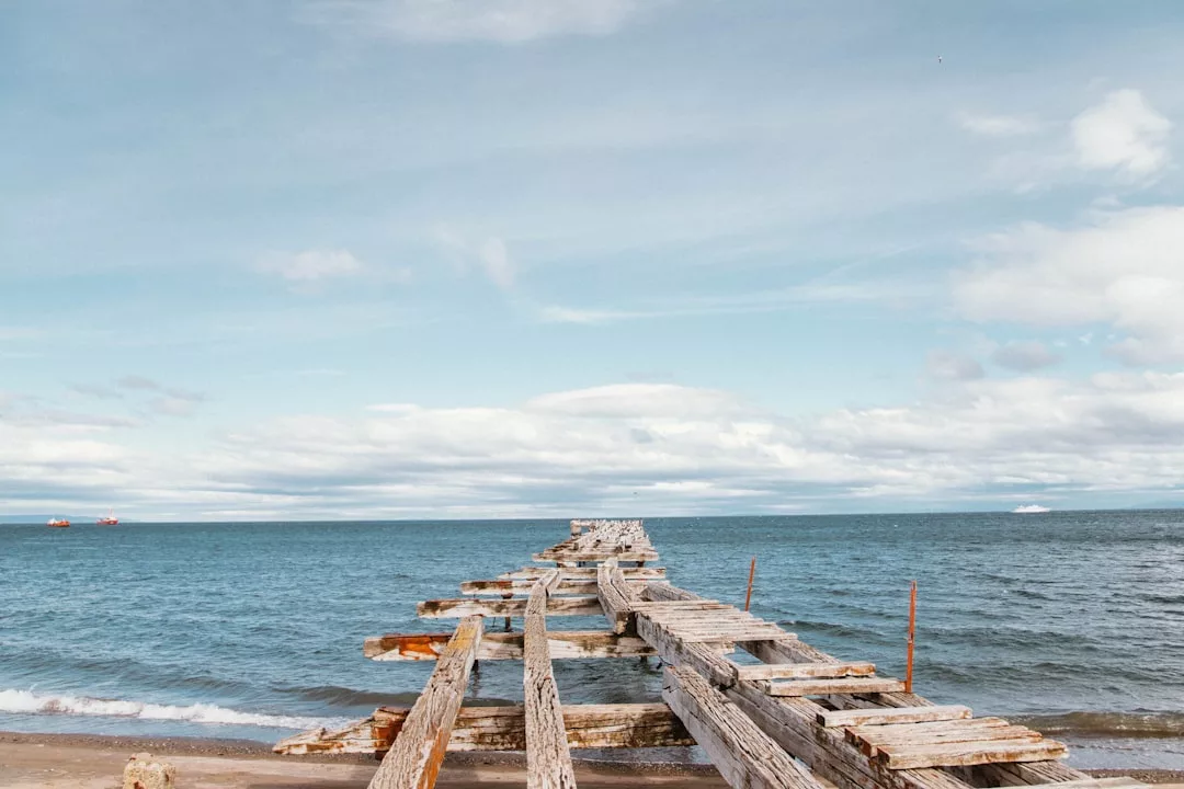 A weathered, broken wooden pier extends into the ocean under a cloudy blue sky, capturing a tranquil coastal scene ideal for real estate with waterfront potential.